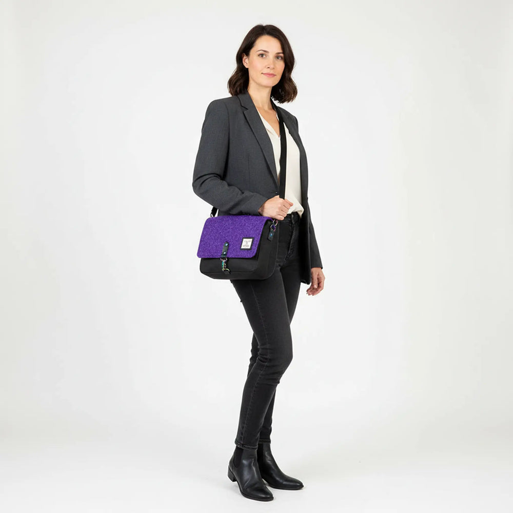 Woman standing in a white studio wearing a grey blazer, cream blouse and black jeans, carrying a black crossbody bag with a purple Harris Tweed® flap