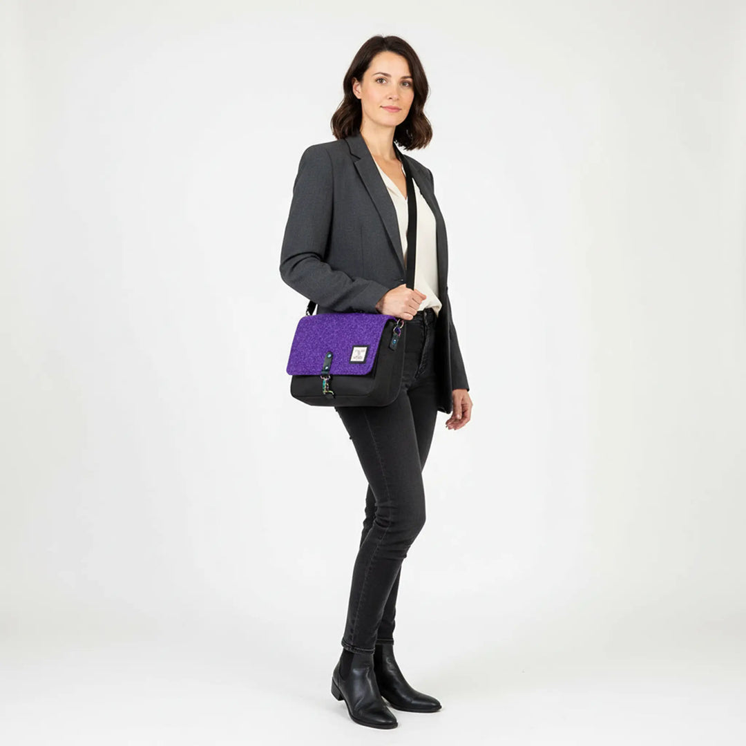 Woman standing in a white studio wearing a grey blazer, cream blouse and black jeans, carrying a black crossbody bag with a purple Harris Tweed® flap