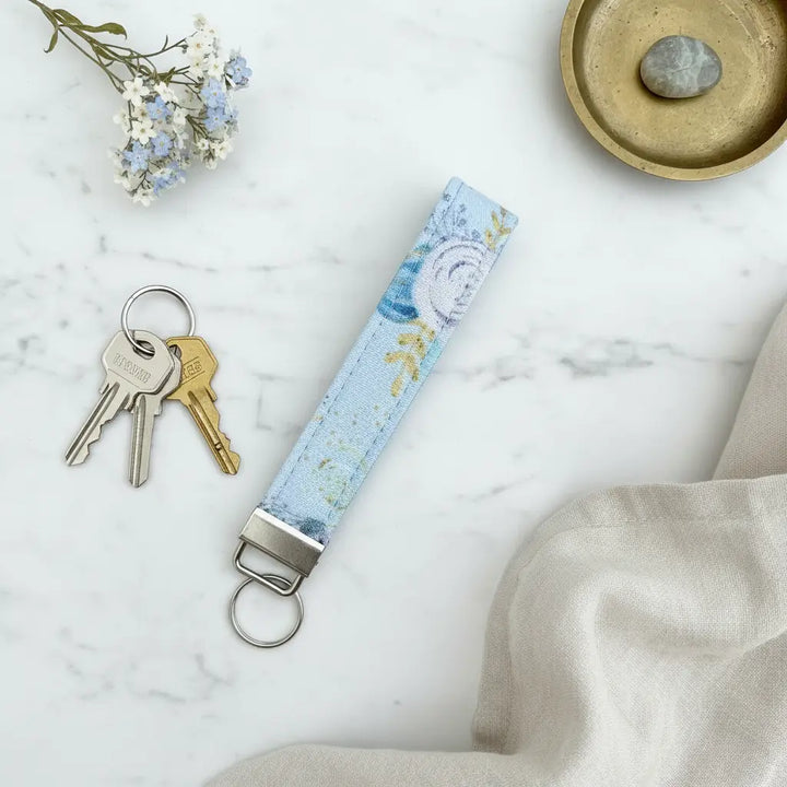 A pale blue floral fabric keyfob with a silver keyring, styled flat on a marble surface beside a set of keys, a brass dish with a grey stone, a sprig of blue and white flowers, and a soft beige linen cloth.