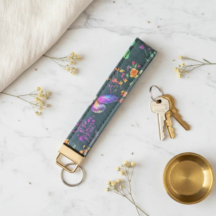 A fabric keyfob featuring a woodland-inspired floral and butterfly print with gold hardware, styled flat on a marble surface beside a set of keys, scattered dried flowers, a brass dish, and a soft cream linen cloth.