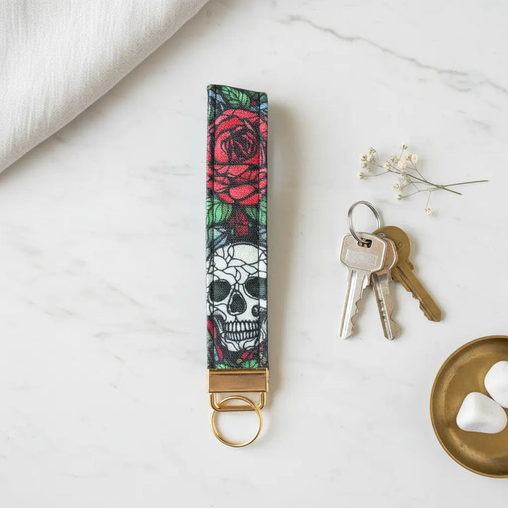 A fabric keyfob with a bold skull and red rose design, finished with gold hardware, styled flat on a marble surface beside a set of keys, a brass dish with white stones, a sprig of delicate flowers, and a soft linen cloth.