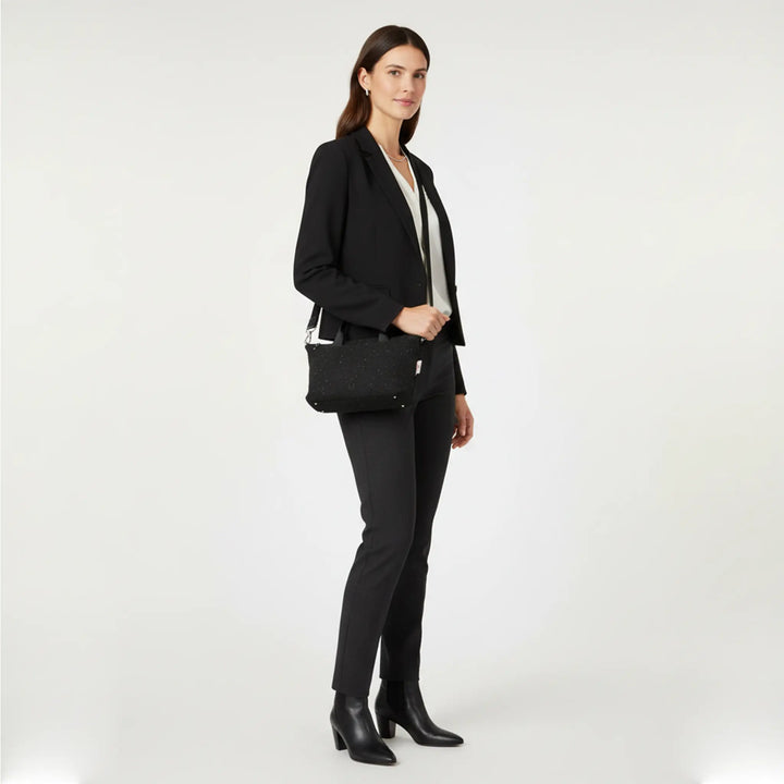 Woman standing in a white studio wearing a black blazer, cream blouse and tailored trousers, holding a small black Harris Tweed® mini tote bag with white nepps worn crossbody