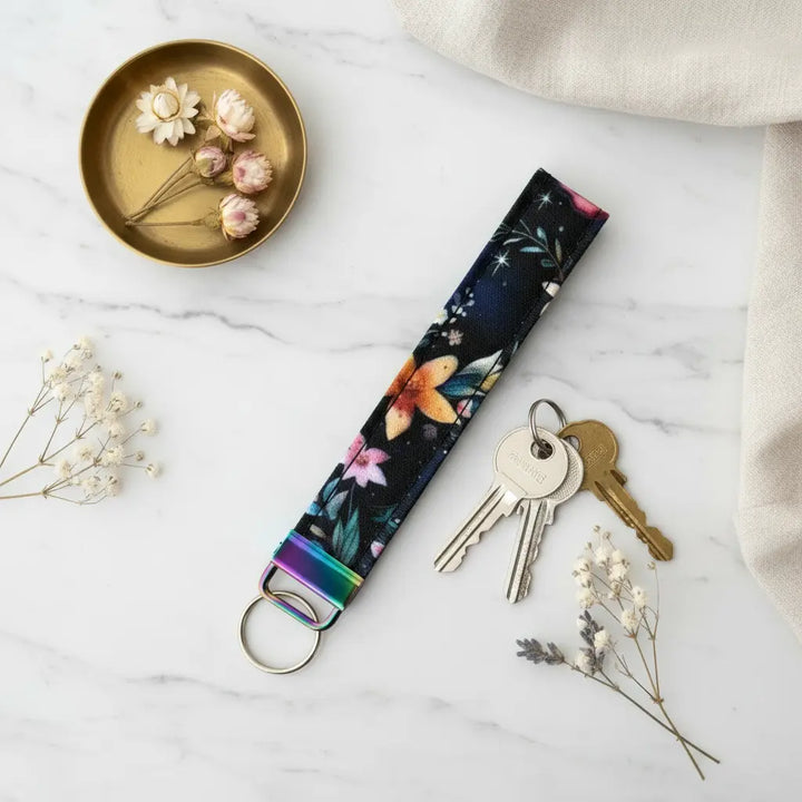 A fabric keyfob with a vibrant floral pattern on a dark background and iridescent metal hardware, styled flat on a marble surface beside a set of keys, dried flowers in a brass dish, and a soft beige linen cloth.