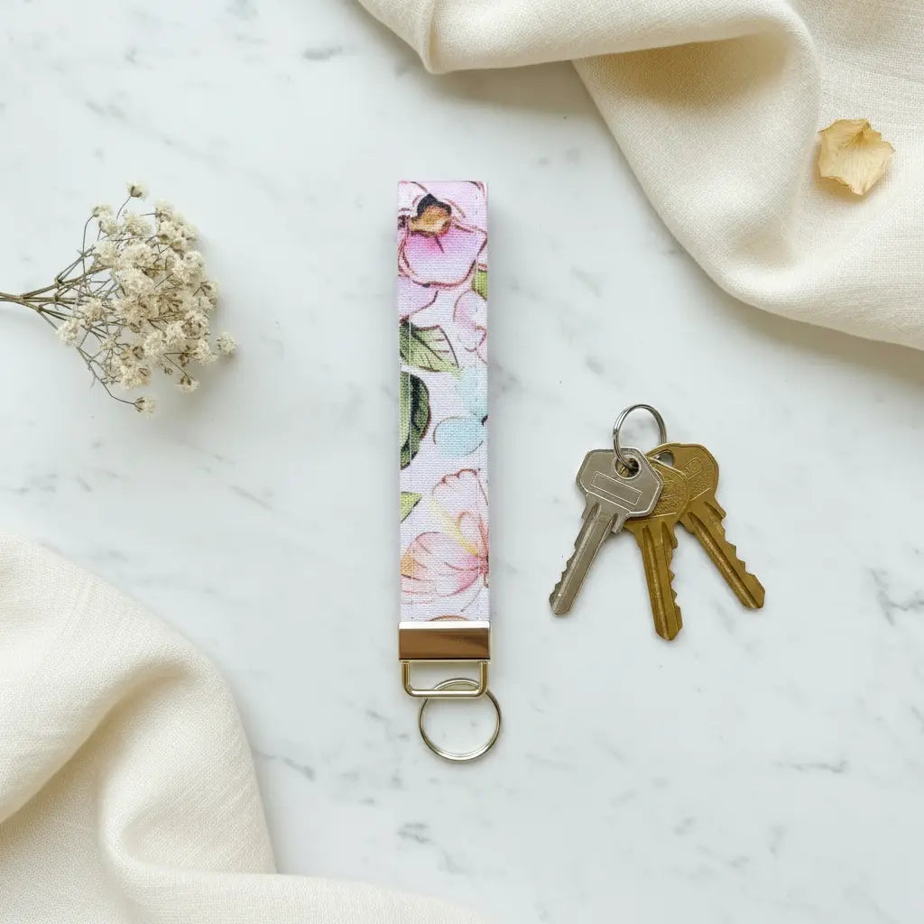 A fabric keyfob with a delicate pastel floral pattern and gold hardware, styled flat on a marble surface beside a set of keys, dried flowers, and soft cream linen cloth.