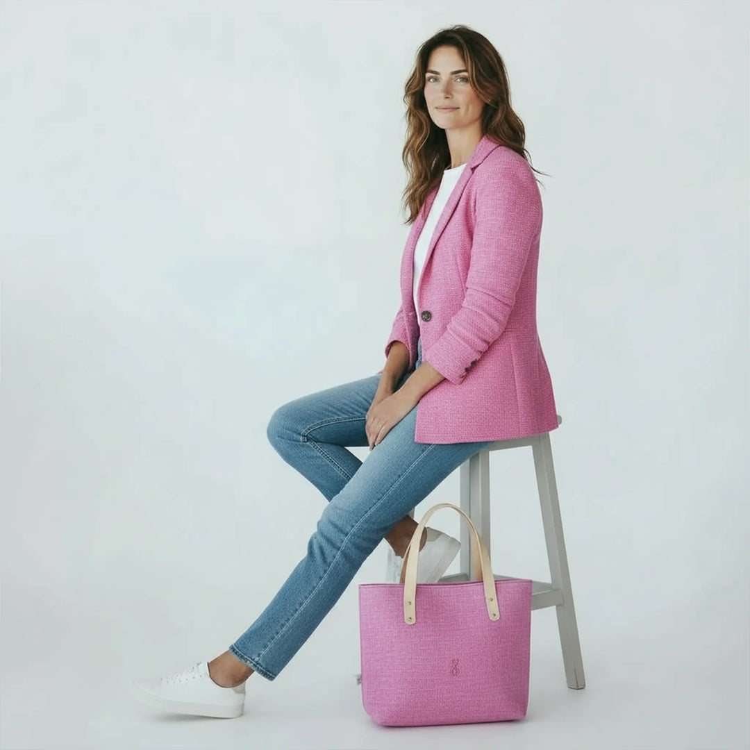 Woman sitting on a stool in a bright white studio wearing a pink blazer and jeans, with a large pink Harris Tweed® tote bag by her feet