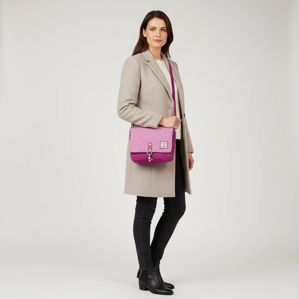 Woman standing in a white studio wearing a long beige coat, black jeans and ankle boots, carrying a pink crossbody bag with a pink Harris Tweed® flap