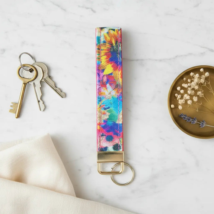 A fabric keyfob with a colourful sunflower and abstract floral print, finished with gold hardware, styled flat on a marble surface beside a set of keys, a brass dish with dried flowers, and a soft cream linen cloth.
