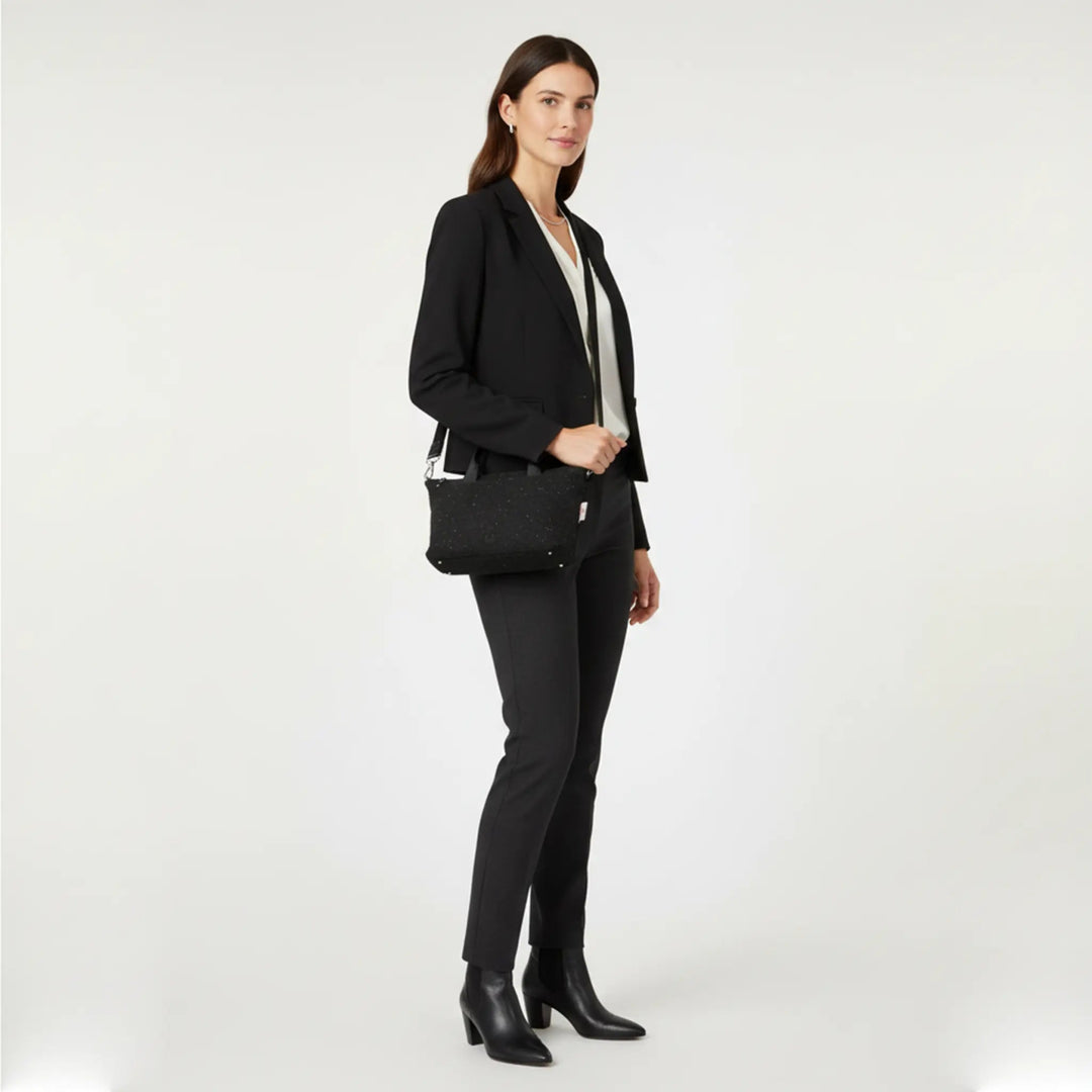 Woman standing in a white studio wearing a black blazer, cream blouse and tailored trousers, holding a small black Harris Tweed® mini tote bag with white nepps worn crossbody