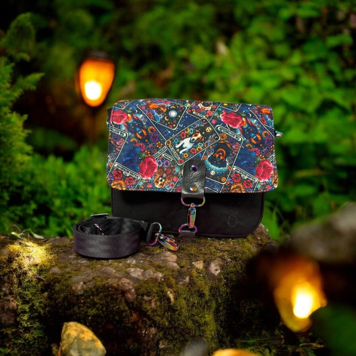 Artisan-made crossbody bag with a tarot card-themed dog print flap, set on a mossy forest rock with glowing lanterns. Features oilskin body and rainbow hardware.