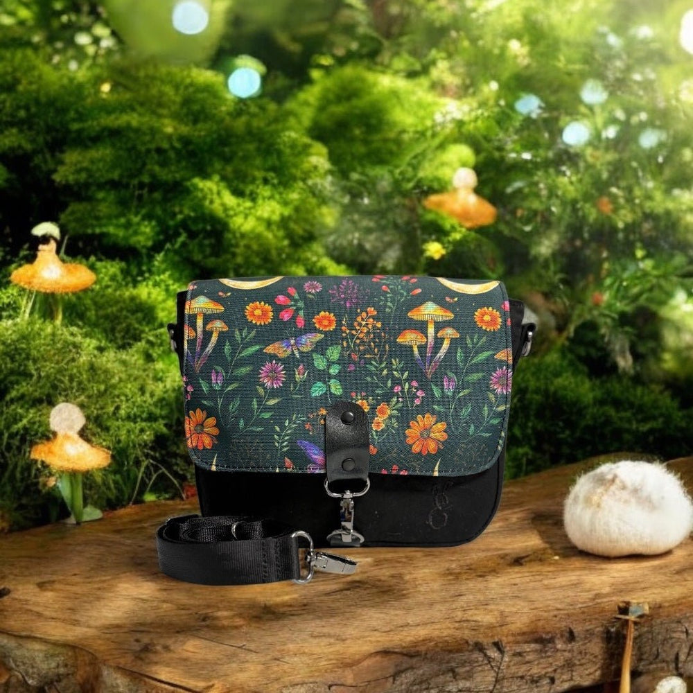 Enchanted Woodland Crossbody Bag displayed in a fairytale-inspired forest setting, surrounded by mossy textures, mushrooms, and glowing orbs. The flap features a whimsical nature print with wildflowers and mushrooms, capturing the magic of the woods.

