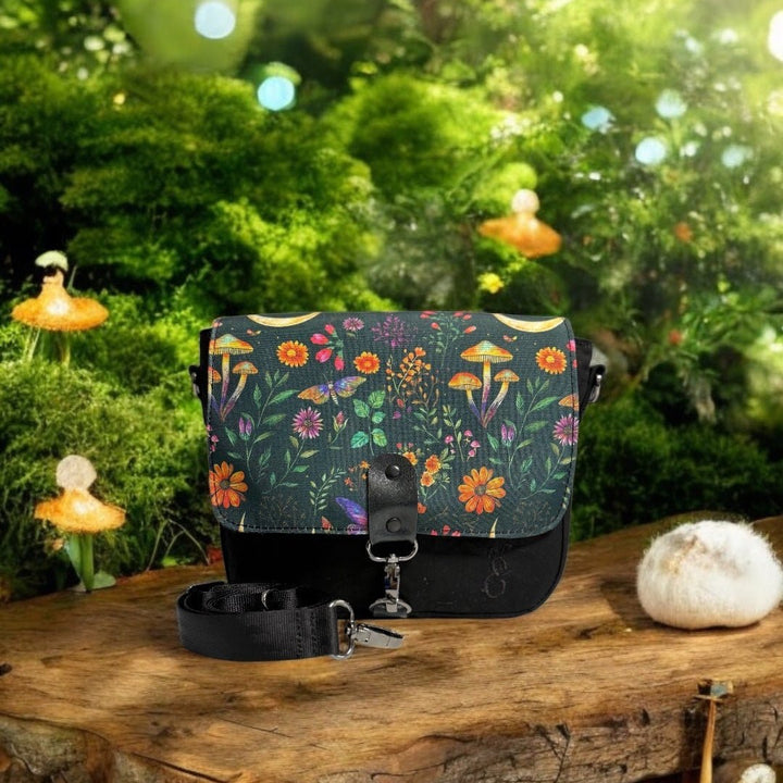 Enchanted Woodland Crossbody Bag displayed in a fairytale-inspired forest setting, surrounded by mossy textures, mushrooms, and glowing orbs. The flap features a whimsical nature print with wildflowers and mushrooms, capturing the magic of the woods.
