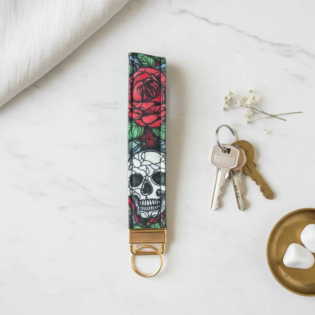 A fabric keyfob with a bold skull and red rose design, finished with gold hardware, styled flat on a marble surface beside a set of keys, a brass dish with white stones, a sprig of delicate flowers, and a soft linen cloth.