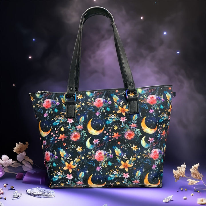 Handcrafted Mystic Garden Tote Bag featuring celestial moons and floral pattern on black background with pink roses, colorful flowers, and golden crescent moons. Water-resistant canvas handbag made in Wales with black  handles.