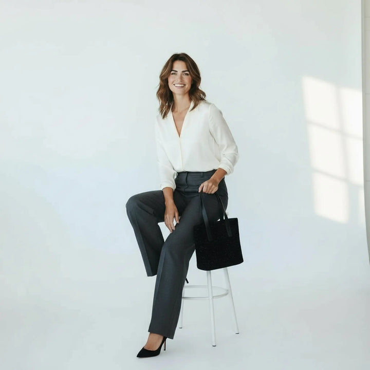 Woman sitting on a stool  in a bright white studio wearing a cream blouse and tailored trousers, holding a black Harris Tweed® tote bag with white nepps in her hand