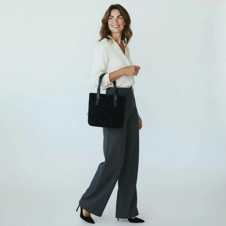 Woman walking in a bright white studio wearing a cream blouse and tailored trousers, carrying a small black Harris Tweed® tote bag with white nepps on her forearm
