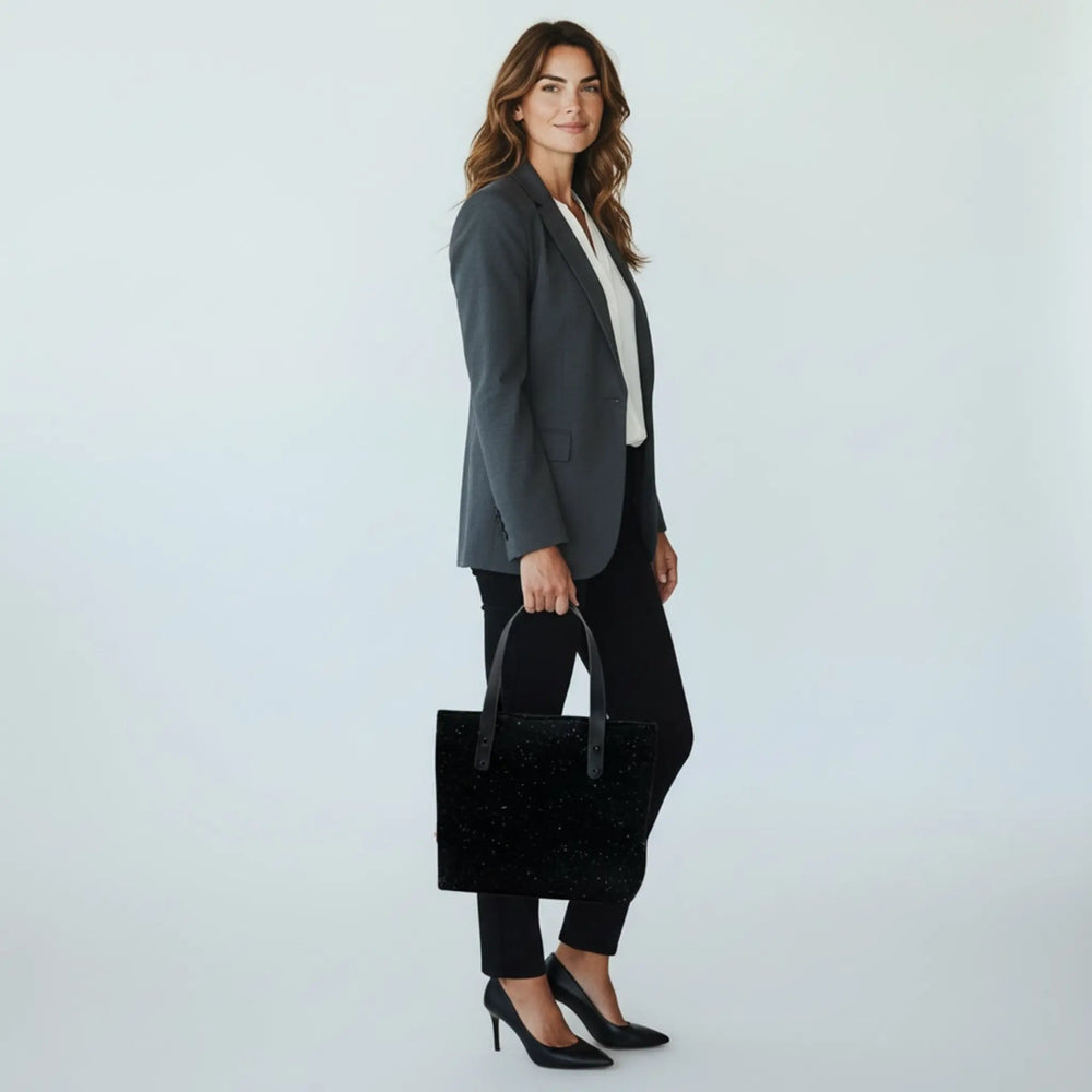 Woman in a bright white studio wearing a grey blazer, black trousers, carrying a black Harris Tweed® tote bag in her hand.