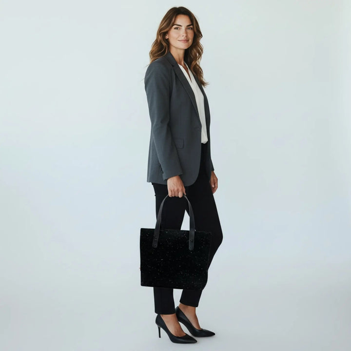 Woman in a bright white studio wearing a grey blazer, black trousers, carrying a black Harris Tweed® tote bag in her hand.