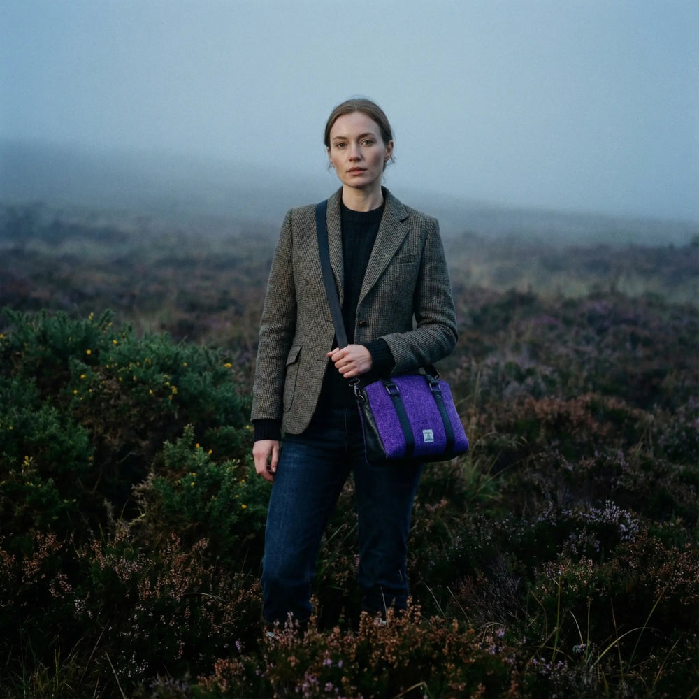Woman standing in misty heather moorland wearing a tweed blazer and jeans, carrying a purple Harris Tweed® tote bag with black leather trim, photographed in a natural British countryside setting.