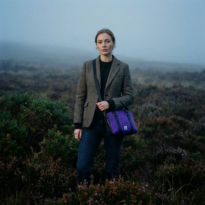 Woman standing in misty heather moorland wearing a tweed blazer and jeans, carrying a purple Harris Tweed® tote bag with black leather trim, photographed in a natural British countryside setting.