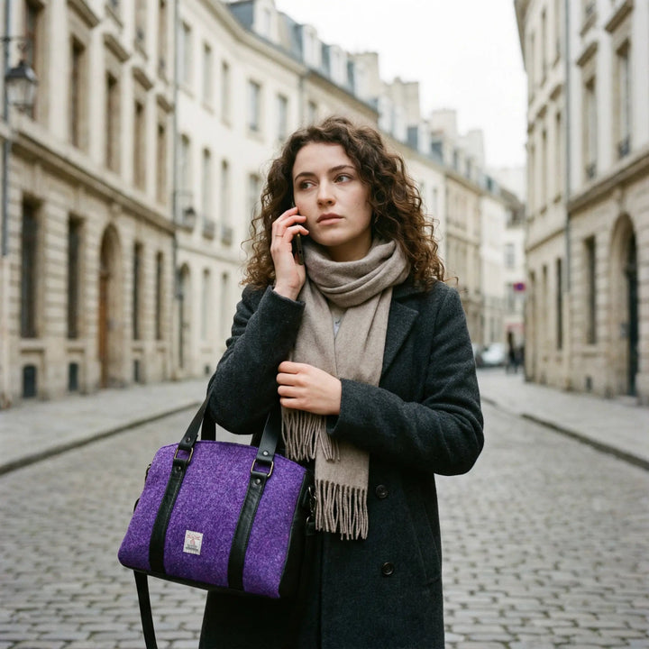 Young woman with curly brunette hair wearing a charcoal wool coat and beige cashmere scarf, carrying a purple Harris Tweed® tote bag with black leather trim, talking on the phone in a classic European city street.